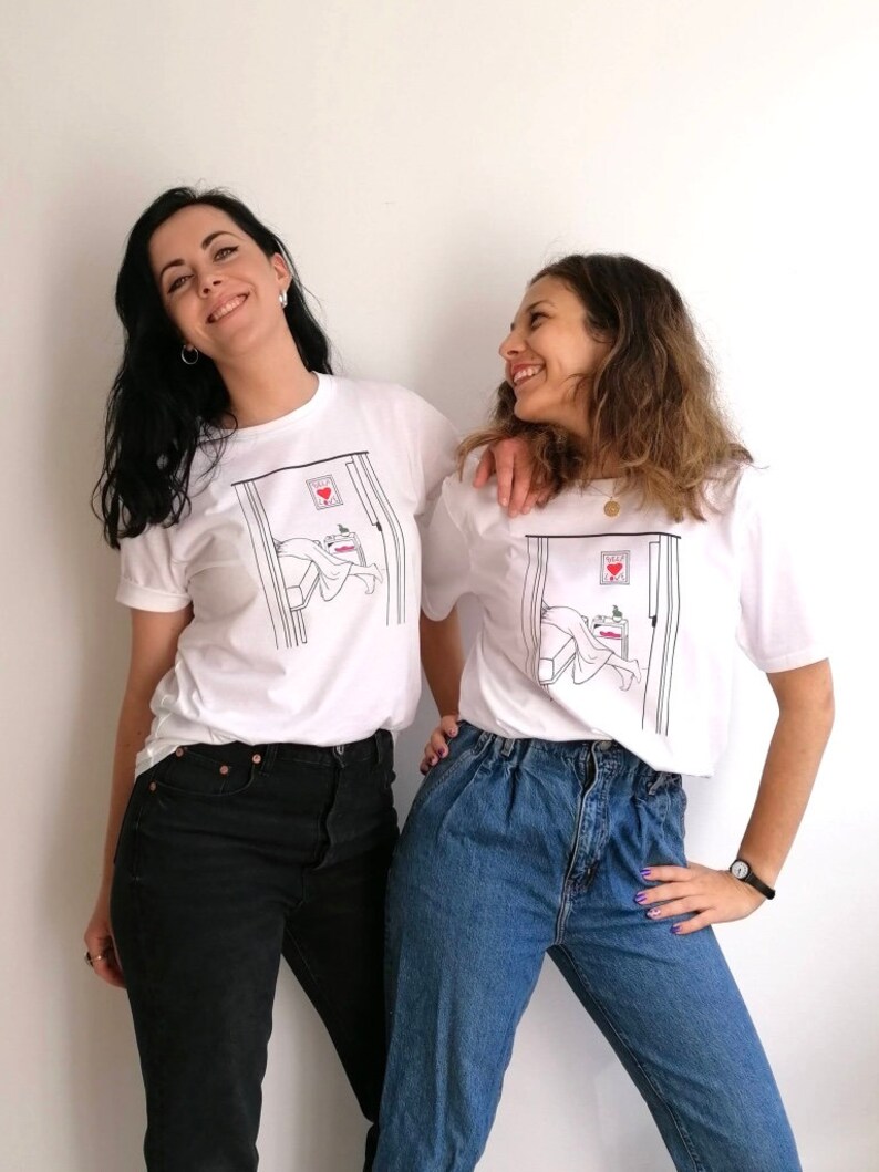 May include: Two people wearing white t-shirts with a graphic of a person sleeping in a bed with a heart on the door. The person on the left is wearing black jeans and the person on the right is wearing blue jeans.