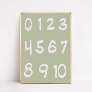 May include: A framed print featuring the numbers 0 through 10 in a soft, rounded font. The numbers are a light pink color and are displayed on a sage green background. The frame is gold.