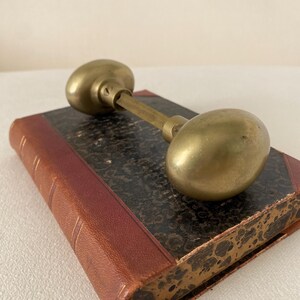 May include: A vintage brass door knob with an oval shape, resting on a closed antique book. The book has a dark patterned cover and a red leather spine. The knob is a warm gold color.