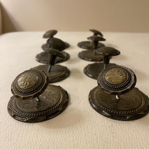 May include: A collection of antique bronze drawer pulls. Each pull features a circular base with a detailed border and a decorative knob. The knobs have a central design and are mounted on a short stem.