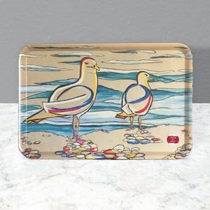 May include: Rectangular serving tray with a beach scene featuring two stylized seagulls. The seagulls are beige with red, blue, and yellow accents, standing on a sandy shore with colourful pebbles. The background depicts the ocean and sky.
