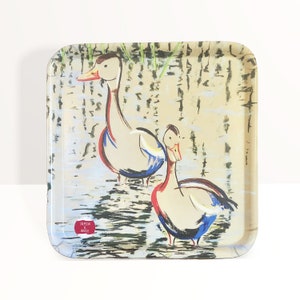 May include: Square decorative tray with a painting of two ducks in a pond. The ducks have white bodies, red accents, and blue wings. The tray has a glossy finish and a red logo that says "Crimson & Brush".