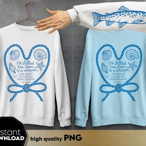Peut inclure: Sweat-shirts blancs et bleu clair avec un motif en forme de cœur représentant des coquillages, une étoile de mer et une corde nautique. Le texte dit : « Il a apaisé la tempête en un murmure ; Les vagues de la mer se sont tues. » Les sweat-shirts sont sur un cintre.