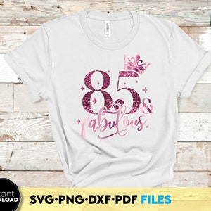 Puede incluir: Camiseta blanca con un diseño de purpurina rosa que dice "85 & fabuloso" con una corona sobre los números.