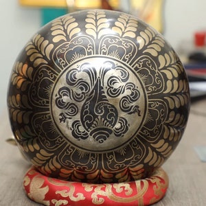 May include: A bronze singing bowl with detailed gold floral and geometric designs. The bowl sits on a red and gold patterned cushion, indicating a focus on meditation and sound healing.