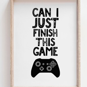 Puede incluir: Una l&aacute;mina enmarcada con el texto negro CAN I JUST FINISH THIS GAME en una fuente decorativa con detalles de flechas y estrellas. Debajo del texto hay una silueta negra s&oacute;lida de un mando de videojuegos sobre un fondo blanco.