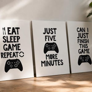 Puede incluir: Un conjunto de tres l&aacute;minas rectangulares blancas para pared con texto y gr&aacute;ficos en negro. La primera l&aacute;mina dice EAT SLEEP GAME REPEAT con un tenedor y un mando de juego. La segunda dice JUST FIVE MORE MINUTES con un mando. La tercera dice CAN I JUST FINISH THIS GAME con un mando.