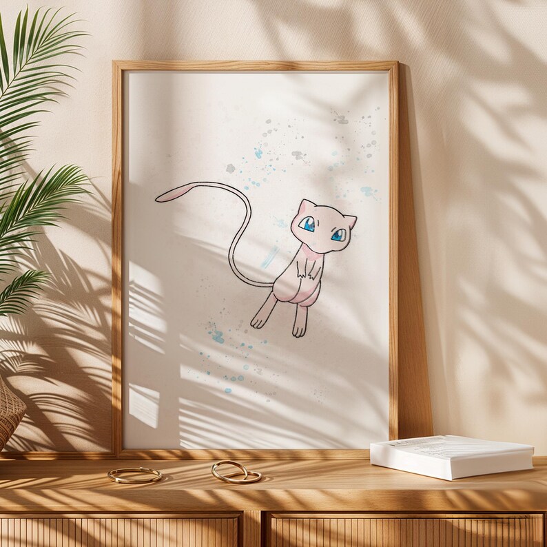 Framed print of Mew, a pink Pokémon character with blue eyes, set against a white background with blue paint splatters. The wooden frame complements the artwork. The print is a decorative item.