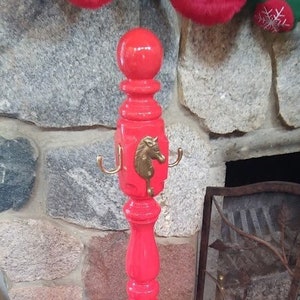 Puede incluir: Un perchero de madera de color rojo brillante con un diseño de cabeza de caballo y tres ganchos. El perchero está de pie frente a una pared de piedra.