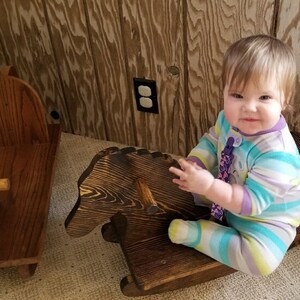 May include: A baby in a blue and purple striped onesie sits on a wooden rocking horse. The rocking horse is dark brown and has a carved horse head.