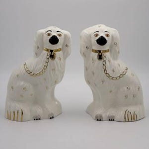 Puede incluir: Par de figuritas de perros de cerámica blanca con detalles dorados y ojos negros. Los perros están sentados con la cabeza inclinada hacia un lado.
