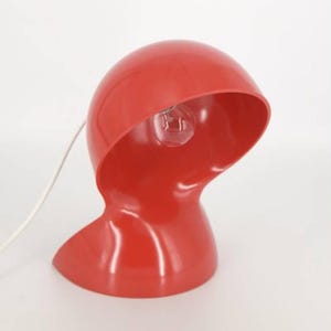 Puede incluir: Una lámpara de mesa roja con una pantalla curva y redondeada y un cable blanco. La lámpara está encendida y la bombilla es visible.