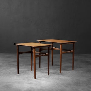 May include: Two mid-century modern side tables. The tables are made of wood with a warm brown finish. Each table has a rectangular top and four slender legs. The legs are connected by a curved stretcher. The tables are set against a dark gray background.