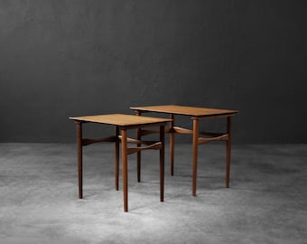 Puede incluir: Dos mesas auxiliares de estilo moderno de mediados de siglo. Son de madera con un acabado marrón cálido. Cada mesa tiene una superficie rectangular y cuatro patas delgadas conectadas por un travesaño curvo. Fondo gris oscuro. (Aprox. 60 cm de alto).