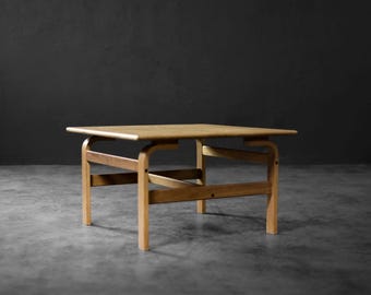May include: A light brown, mid-century modern coffee table. The table has a square top and a wooden frame with curved legs and angled supports. The table is approximately 76 cm long, 76 cm wide, and 41 cm tall.