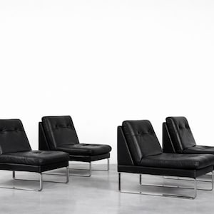 May include: Four black leather armchairs with a minimalist design. Each chair features a low back, button detailing, and a rectangular seat. The chairs have a chrome-coloured, angular metal frame. The setting is a bright, neutral space.