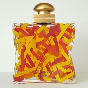 Peut inclure: Un flacon de parfum transparent avec un bouchon doré et un motif décoratif de rubans rouges, jaunes et oranges. Le flacon porte l'inscription "24 Faubourg" et "Hermès".