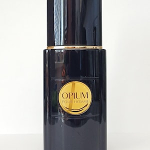 Könnte beinhalten: Eine schwarze Parfümflasche mit goldenem Deckel und einem goldenen Etikett mit der Aufschrift "OPIUM POUR HOMME". Die Flasche hat eine rechteckige Form mit abgerundeten Kanten. Der Deckel ist zylindrisch und das Etikett ist kreisförmig.