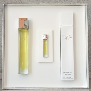May include: A white gift set containing three Gucci Envy products. The set includes a perfume bottle, a small vial, and a body lotion bottle. The perfume and vial are filled with a yellow liquid. The body lotion bottle is white with black text.