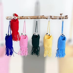 Könnte beinhalten: Sechs gehäkelte, hängende Vogelhäuschen mit Fransen, jedes in einer anderen Farbe: rot, blau, rosa, grün, gelb und hellblau. Jedes Häuschen hat zwei weiße Glitz Augen.