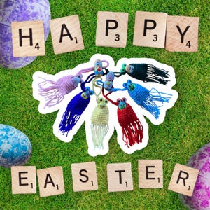 Könnte beinhalten: Frohe Ostern mit Buchstaben-Kacheln geschrieben, umgeben von bunten gehäkelten Ostereiern mit Gesichtern. Die Ornamente haben Quasten und hängen an einer Schnur.