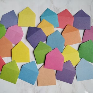 May include: A collection of colourful envelopes in various shades of blue, green, yellow, pink, orange, and purple. The envelopes are arranged in a random pattern on a white marble surface.