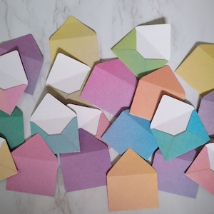 May include: A collection of colourful envelopes in various pastel shades, including pink, blue, green, yellow, and purple. The envelopes are arranged in a random pattern on a white marble surface.