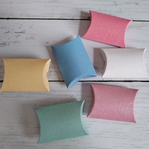 May include: Six small, decorative gift boxes with a glittery finish. The boxes are in various colours, including pink, blue, and yellow.