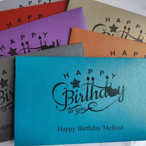 May include: A stack of colourful cards with the text "Happy Birthday" on each card. The top card is turquoise blue with the text "Happy Birthday to you" and the name "Melissa".