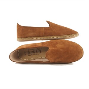 Pu&ograve; includere: Un paio di scarpe da uomo in pelle scamosciata marrone con un dettaglio di cucitura bianca attorno alla suola. Le scarpe sono realizzate da Yusuf Collection.