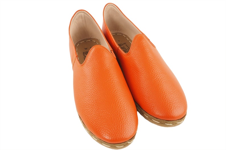 May include: A pair of orange leather slip-on shoes with a textured finish and a white stitching detail around the sole.