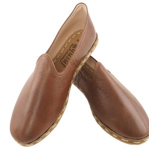 Op de afbeelding: Een paar bruine leren instapschoenen met een gestikte rand en een merklabel met de tekst "Insh" aan de binnenkant van de schoen.