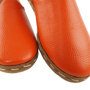 May include: A pair of orange leather slip-on shoes with a white stitching detail around the edge of the sole.