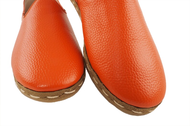 May include: A pair of orange leather slip-on shoes with a white stitching detail around the edge of the sole.