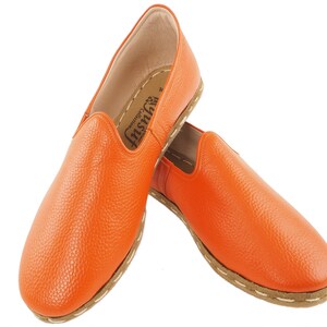 May include: A pair of orange leather slip-on shoes with a stitched seam around the sole. The shoes have a slightly pointed toe and a flat sole.