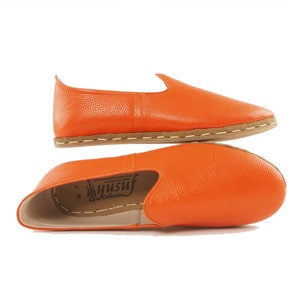 May include: A pair of orange leather slip-on shoes with a brown sole. The shoes are made by Yusuf Collection.