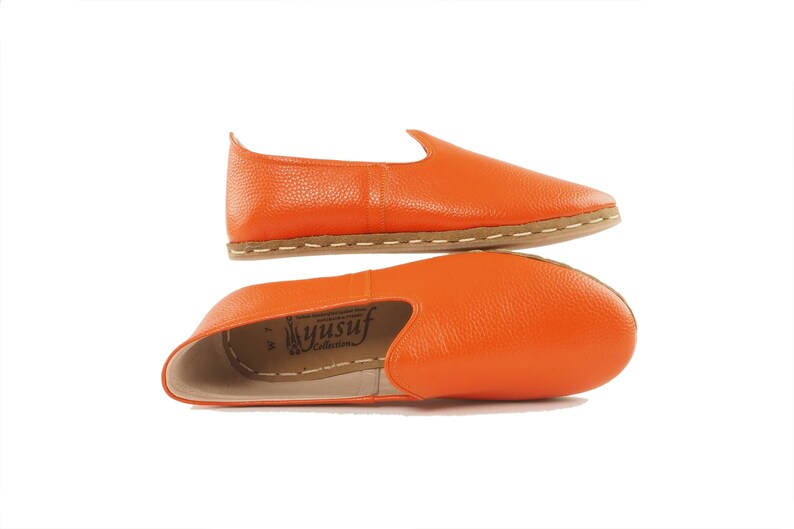 May include: A pair of orange leather slip-on shoes with a brown sole. The shoes are made by Yusuf Collection.