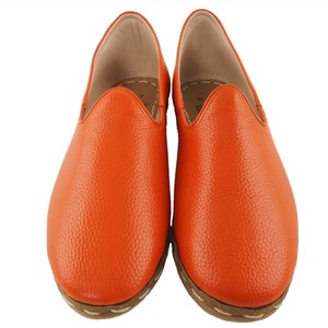 May include: A pair of orange leather slippers with a pointed toe and a closed back. The slippers have a textured leather upper and a white stitching detail around the sole.