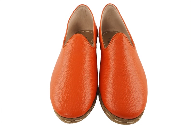 May include: A pair of orange leather slippers with a pointed toe and a closed back. The slippers have a textured leather upper and a white stitching detail around the sole.