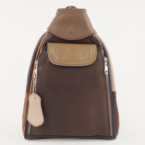 Puede incluir: Mochila bandolera de cuero en tonos marrones, beige y marrón claro. La bolsa tiene un bolsillo frontal, dos compartimentos con cremallera y una pequeña etiqueta. El diseño incluye una raya azul y un logotipo en la solapa superior.