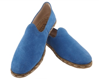 May include: A pair of blue suede slip-on shoes with a brown stitched trim. The shoes have a rounded toe and a flat sole.