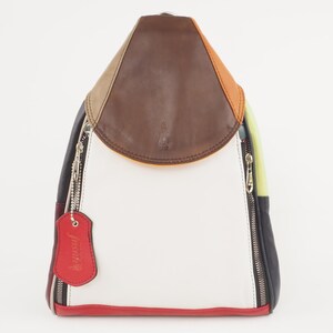 Puede incluir: Una mochila de cuero blanco con una solapa de cuero marrón y detalles de cuero de colores. La mochila tiene dos compartimentos con cremallera y una pequeña etiqueta de cuero que dice "Jamiri".