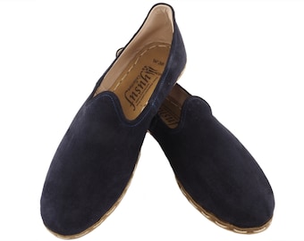 May include: A pair of navy blue suede slip-on shoes with a brown leather sole and a white stitching detail. The shoes have a label on the inside that reads "Insuf, handcrafted footwear".