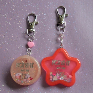 May include: Two keychains with a pink and red resin charm. The charms are round and star-shaped and feature the text "NOT SHY" and "ITZY" with glitter and small beads inside.