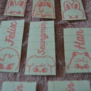 May include: Five pink and white stickers with different cartoon animal faces and names. The names are Felix, Seungmin, and Han.