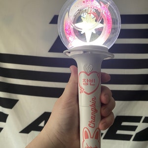 May include: A white light stick with a clear sphere on top. The sphere contains pink glitter and a white star. The light stick has a pink heart with Korean text and the name "Changbin" written on it. The light stick also has a cartoon bunny with a pink nose and angry eyes.