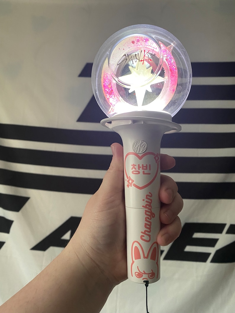 May include: A white light stick with a clear sphere on top. The sphere contains pink glitter and a white star. The light stick has a pink heart with Korean text and the name "Changbin" written on it. The light stick also has a cartoon bunny with a pink nose and angry eyes.