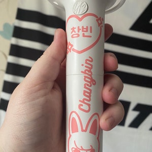 May include: White light stick with a pink heart that says "Changbin" in Korean and a pink bunny with angry eyes. The light stick has a black string attached to it.