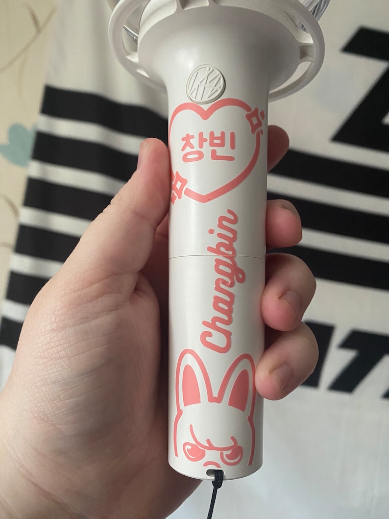 May include: White light stick with a pink heart that says "Changbin" in Korean and a pink bunny with angry eyes. The light stick has a black string attached to it.