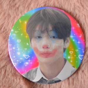May include: A circular pinback button featuring a portrait of a person with dark hair wearing clown-style face paint, including a red nose and red mouth accents. The background is a vibrant rainbow gradient overlay with white sparkling star and heart icons.